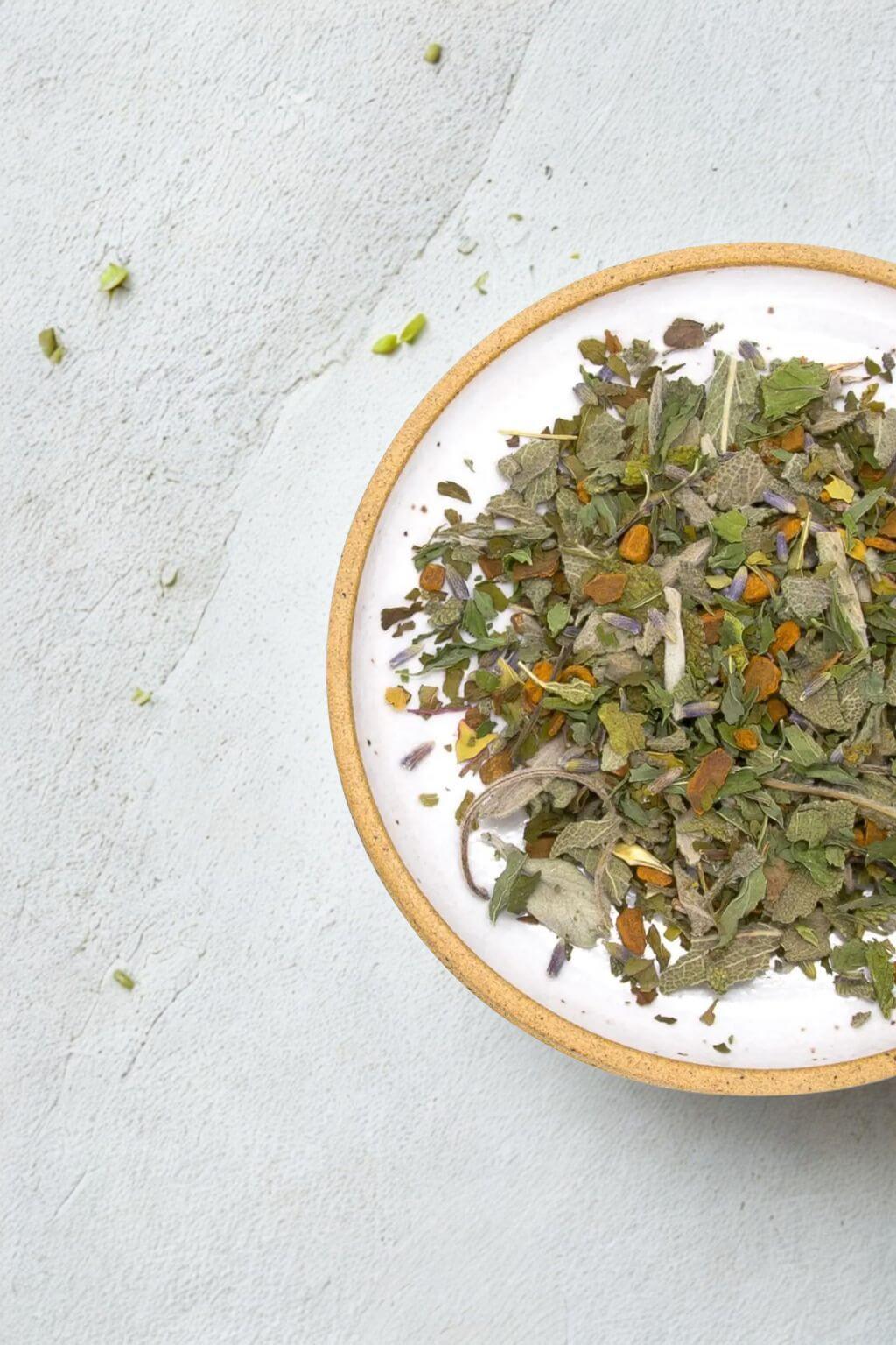 A top-down view of a white ceramic bowl with natural clay rim, filled with a loose herbal tea blend containing green, brown, and orange dried leaves and flowers, set on a textured light gray surface.