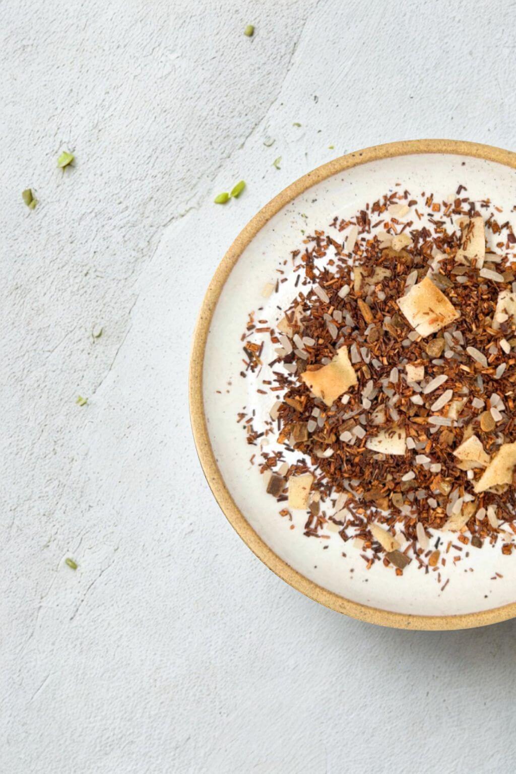 A top-down view of a ceramic bowl filled with a loose herbal tea blend, featuring rooibos, toasted coconut flakes, and rice, set on a light gray textured surface.