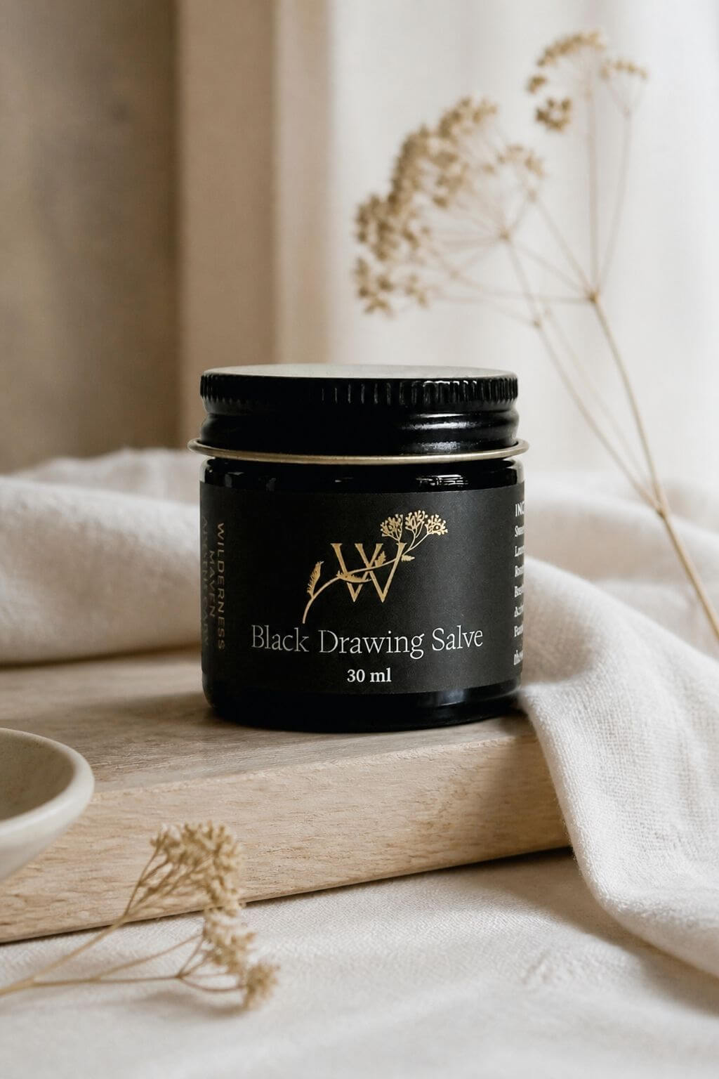 A glass jar of 'Black Drawing Salve' with gold lettering and botanical illustrations, resting on a light wooden board draped with cream-colored fabric and surrounded by dried flowers.