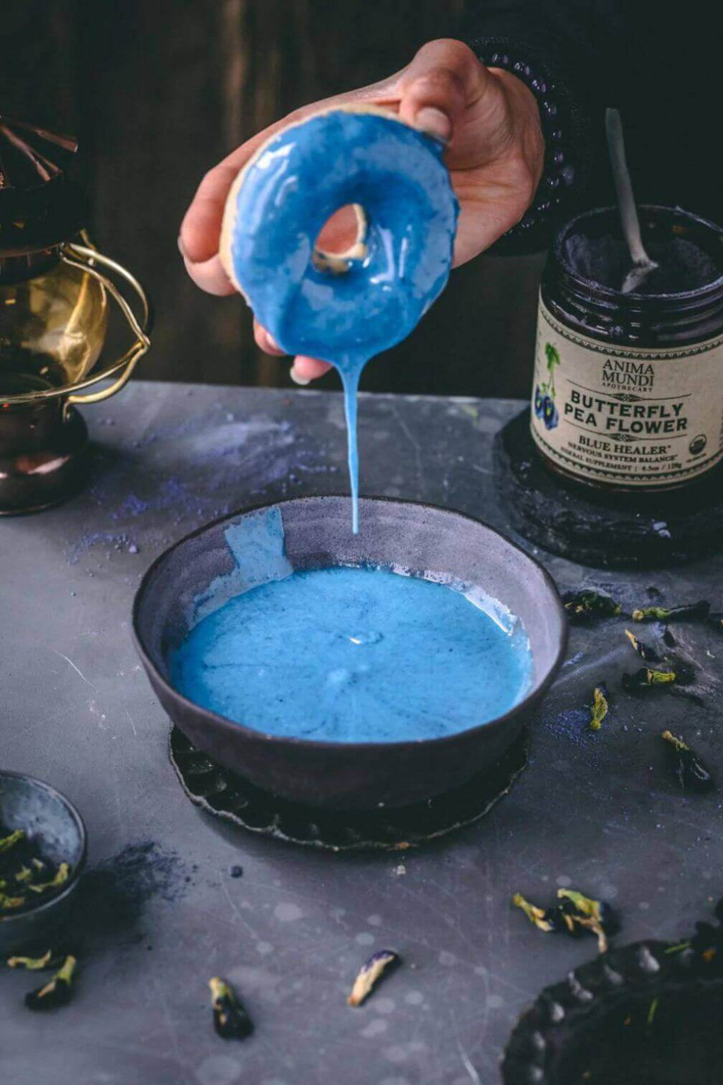 A hand holds a blue glazed donut above a bowl of blue icing, with a jar labeled Butterfly Pea Flower nearby on a dark tabletop.