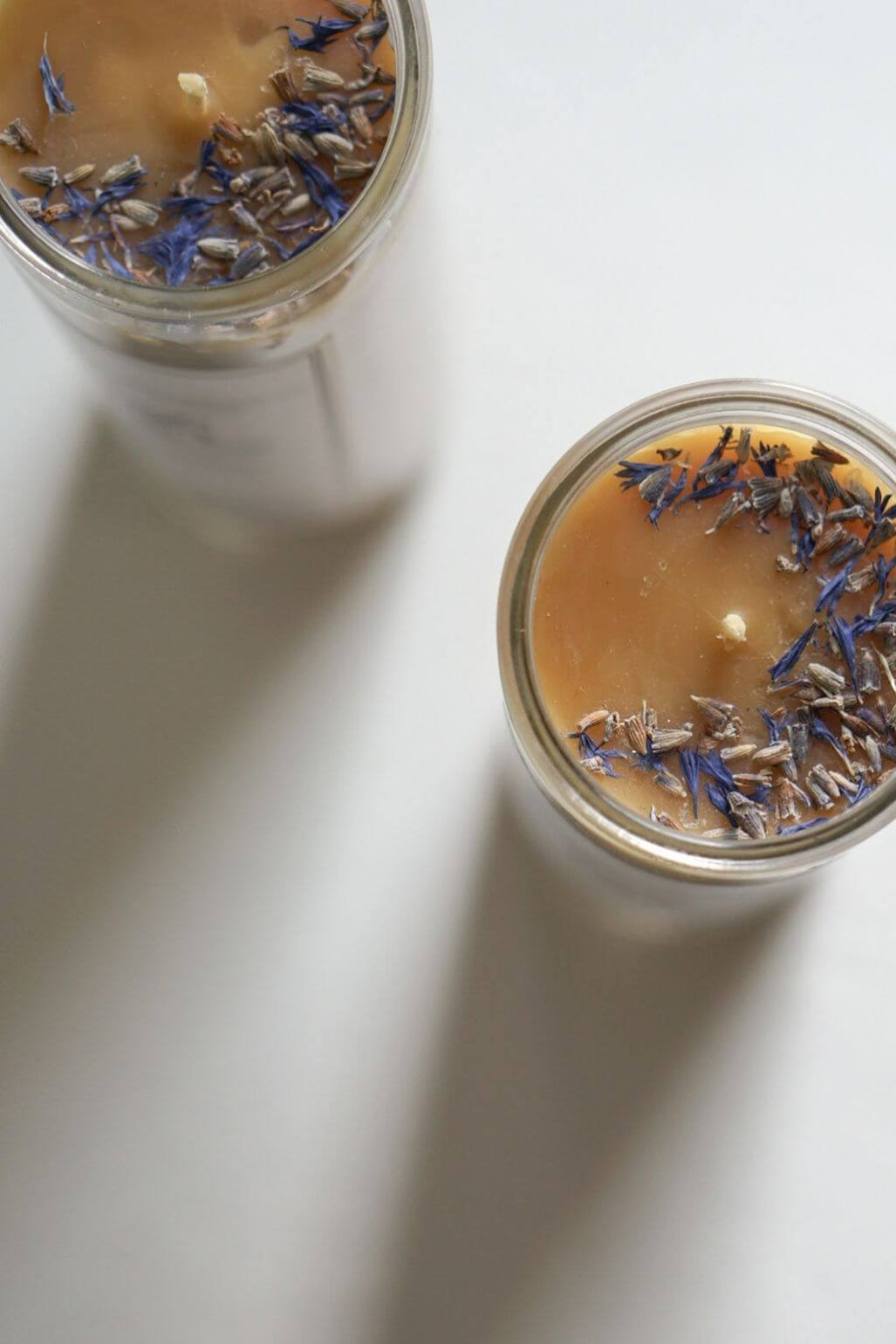 Two clear glass jars containing beeswax candles with dried lavender buds on top, photographed on a clean white surface with soft shadows.