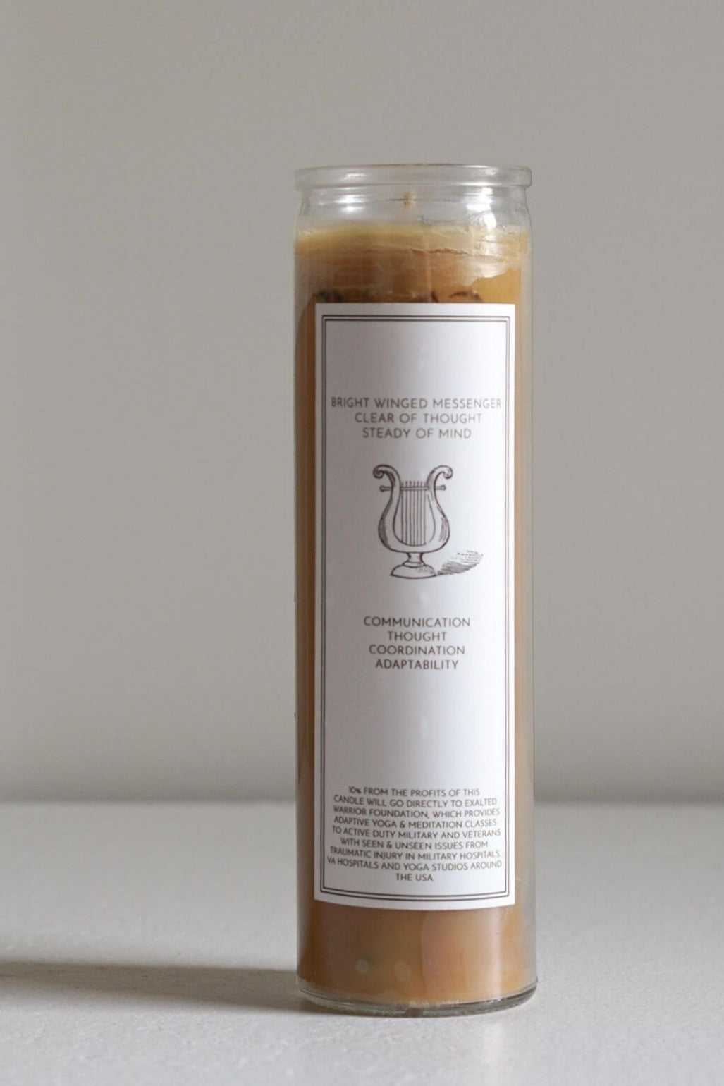 A tall glass jar candle filled with light brown beeswax, featuring a white paper label with black text and a simple lyre illustration, set against a plain light gray background.