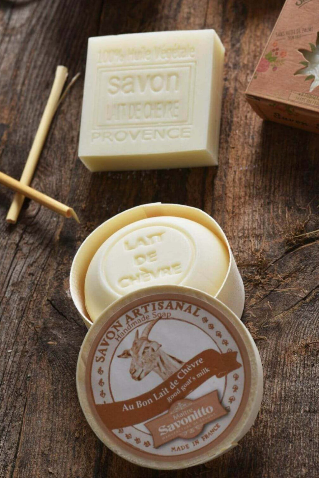 A styled product shot of Savonitto artisanal soap on a rustic wooden surface. The image features an open round tin of 'Lait de Chèvre' (cow's milk) soap showing its pale yellow contents, and the round lid in the foreground with a brown and cream label depicting a goat and the Savonitto brand name.