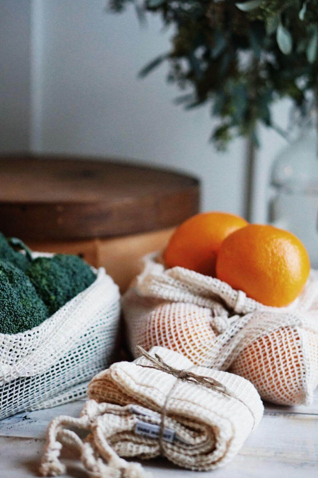 Mesh produce bags with oranges and broccoli on a wooden surface with a plant in the background.