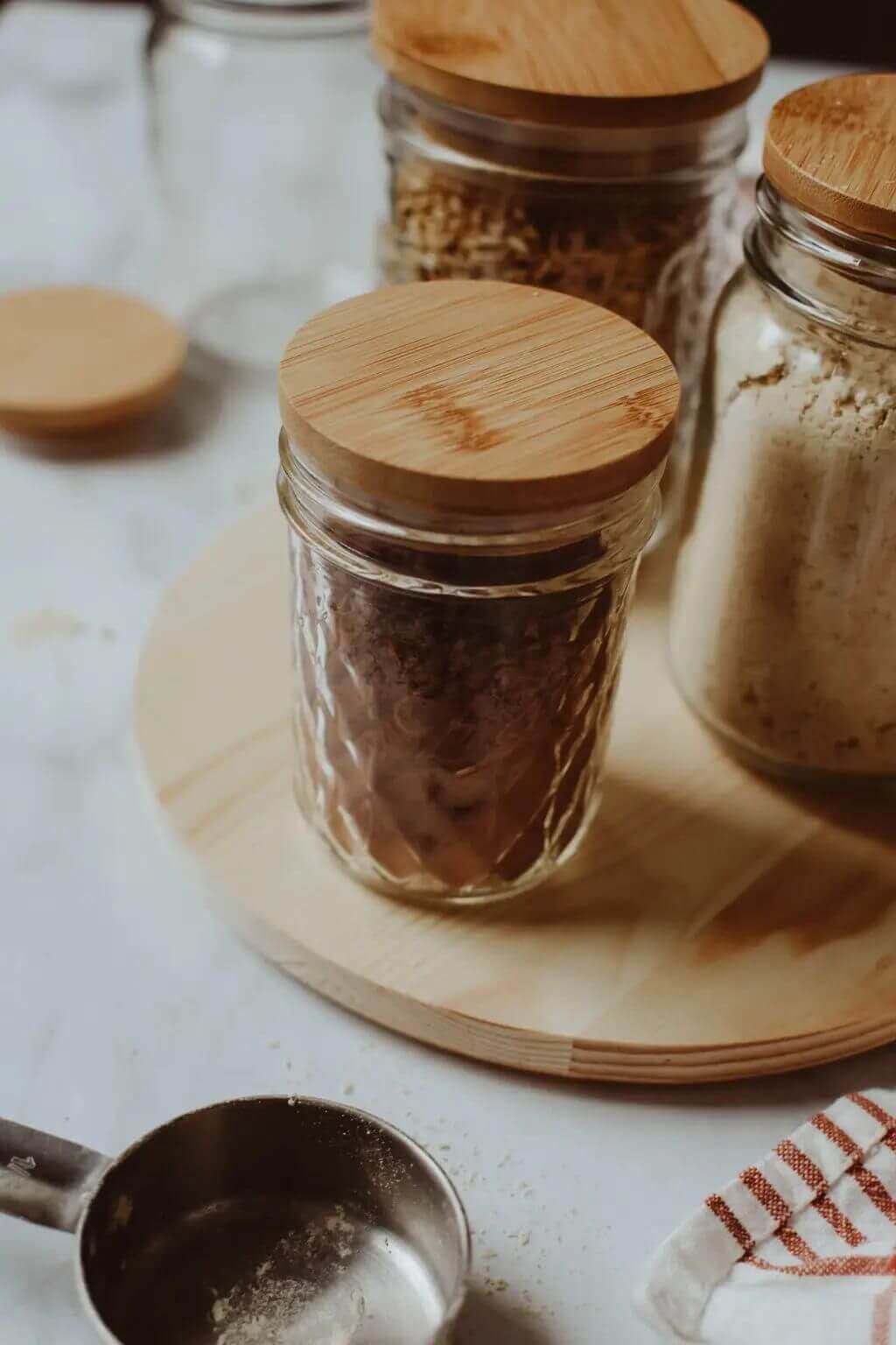 Glass jars with wooden lids on a wooden board with a measuring spoon and towel in the background.