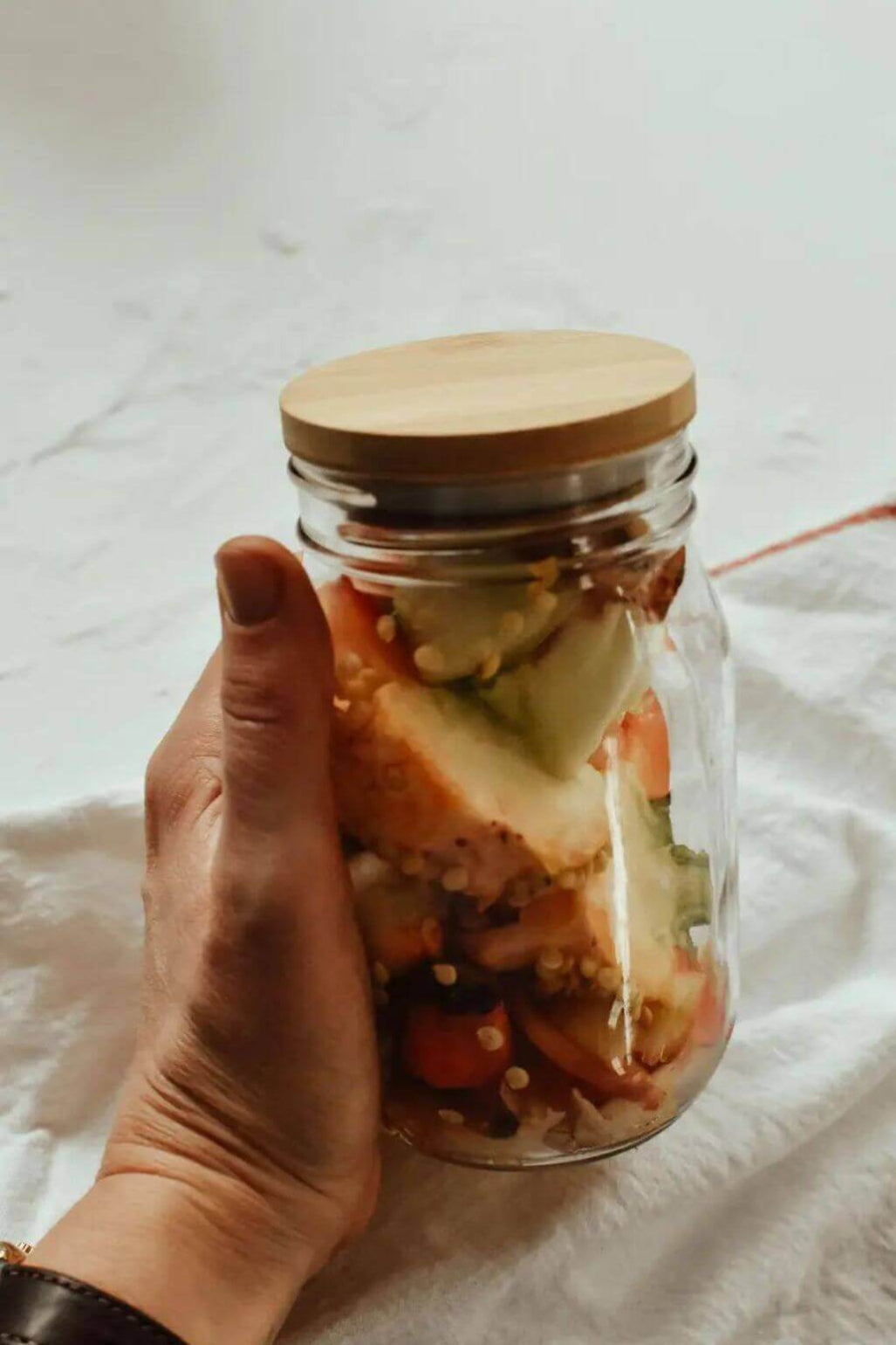 A hand holding a clear glass mason jar with a light bamboo lid set against a white fabric background.
