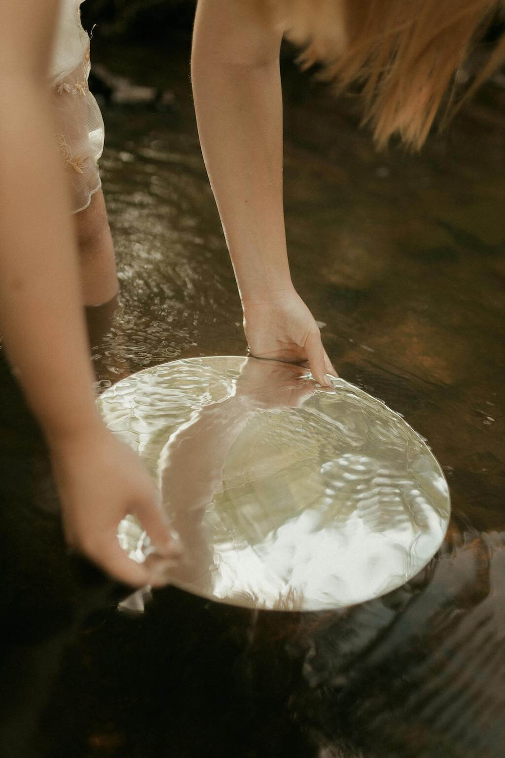 A moody image of a woman holding a mirror into a flowing body of water, reflecting the sky and trees above