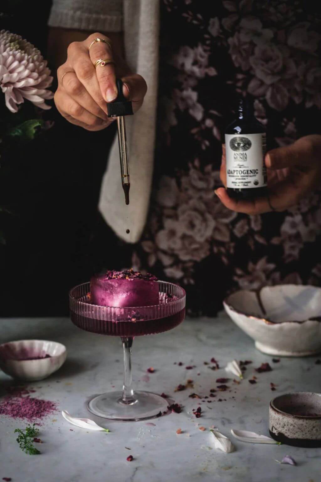 Dropper adding adaptogenic tonic to a vibrant botanical elixir, surrounded by petals and herbs for a moody, ritual-inspired apothecary moment