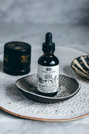 Anma Munde Adaptogenic herbal supplement in an amber glass dropper bottle, styled on a textured ceramic dish with a black container in the background.