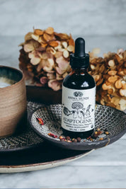A bottle of Anima Mundi Adaptogenic herbal supplement with a black dropper cap and white label, styled on a patterned ceramic dish with dried flowers and a cup on a light marble surface.