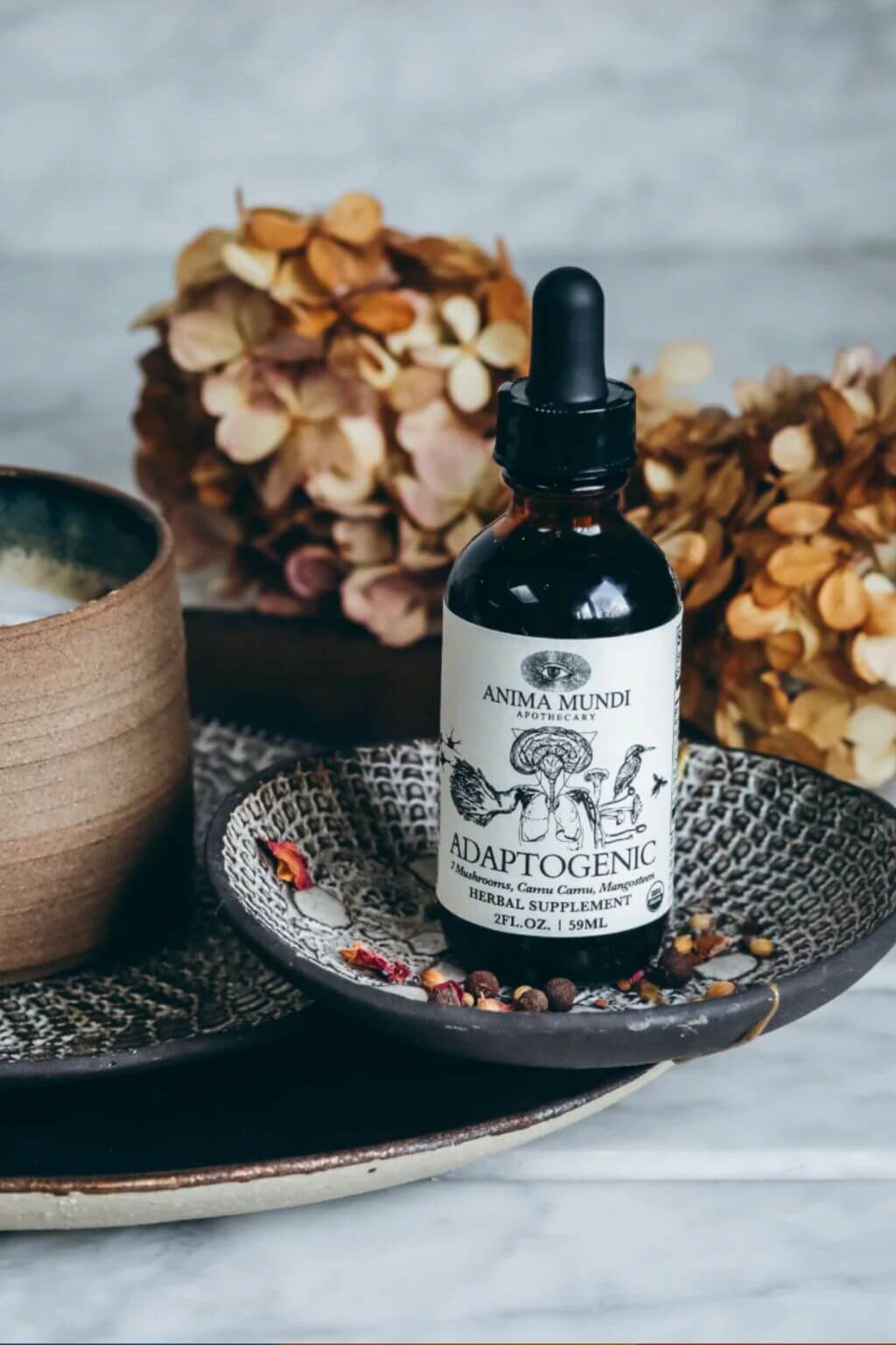 A bottle of Anima Mundi Adaptogenic herbal supplement with a black dropper cap and white label, styled on a patterned ceramic dish with dried flowers and a cup on a light marble surface.