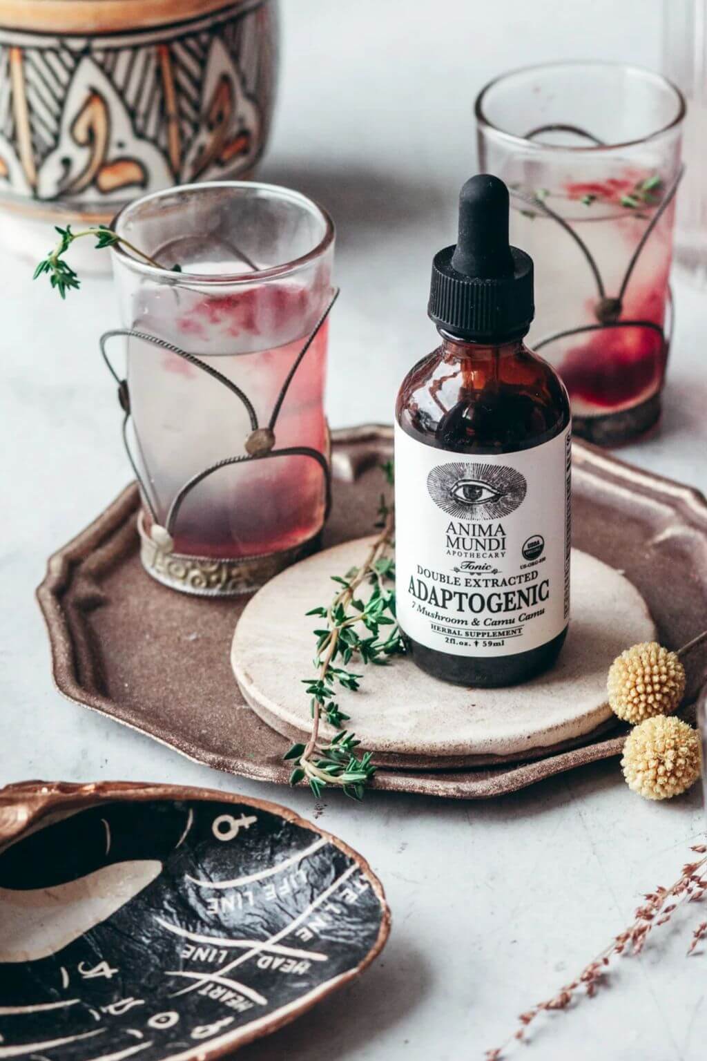 An amber glass dropper bottle labeled Anima Mundi Adaptogenic Herbal Extract with two decorated drinking glasses containing pink drinks, arranged on rustic ceramic plates with herbs and decorative props.