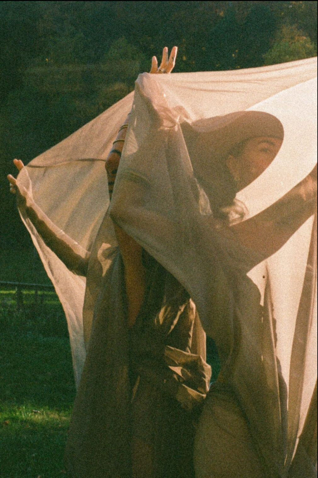 Two women outdoors draped under flowing sheer fabric with arms raised in a natural setting