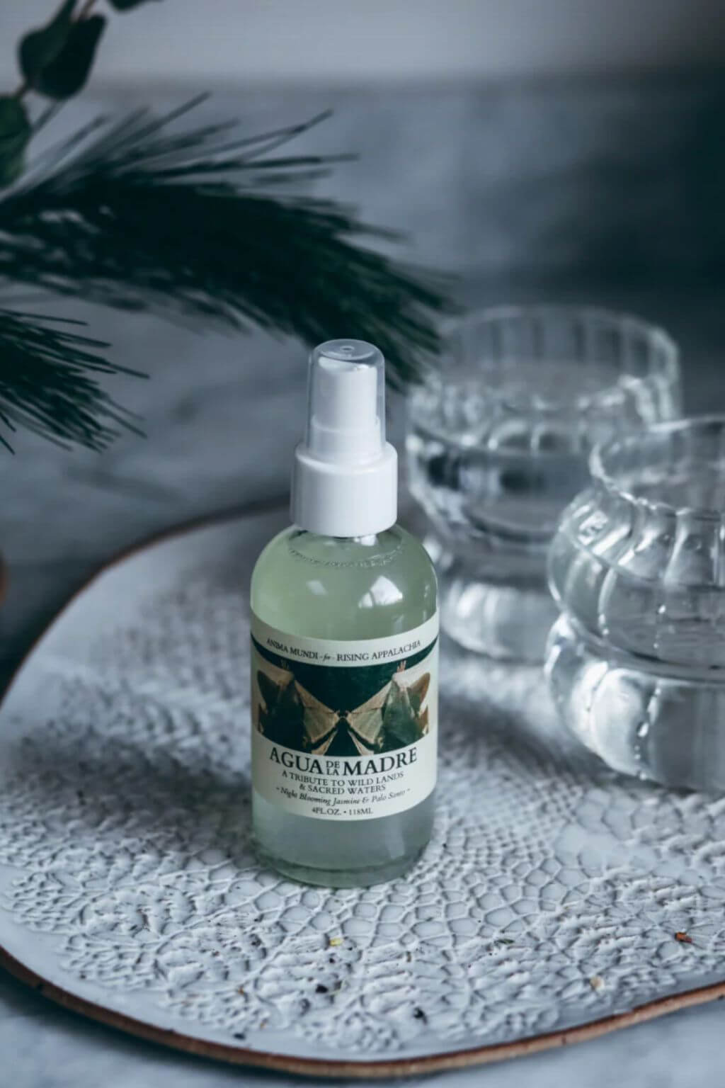 A bottle of Agua de la Madre natural mist with a white spray top, placed on a textured white ceramic plate with glass cups and pine branches in the background.