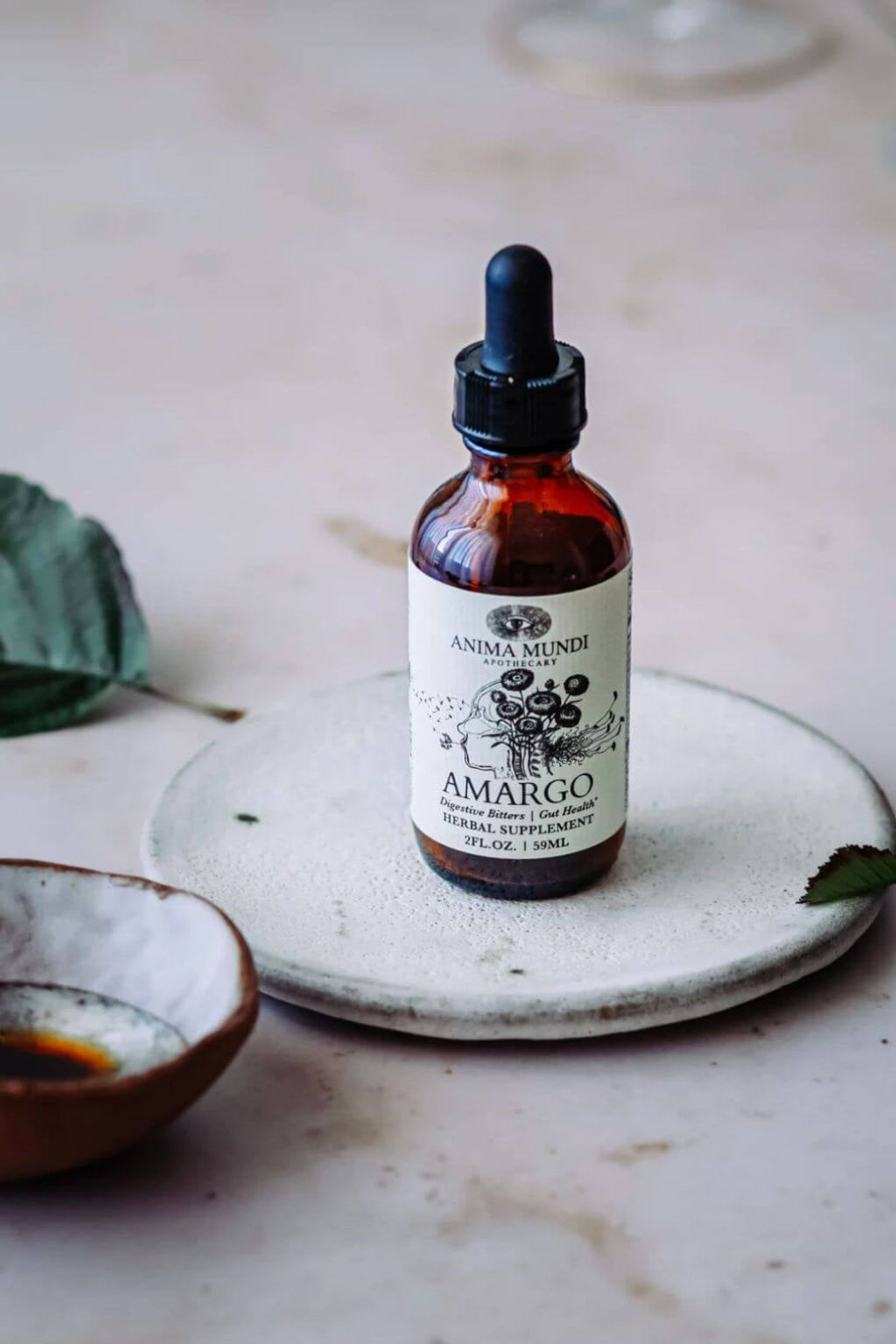 An amber glass dropper bottle labeled Anima Mundi Amargo herbal bitters, placed on a round ceramic dish with a small bowl and leaves on a light stone surface.