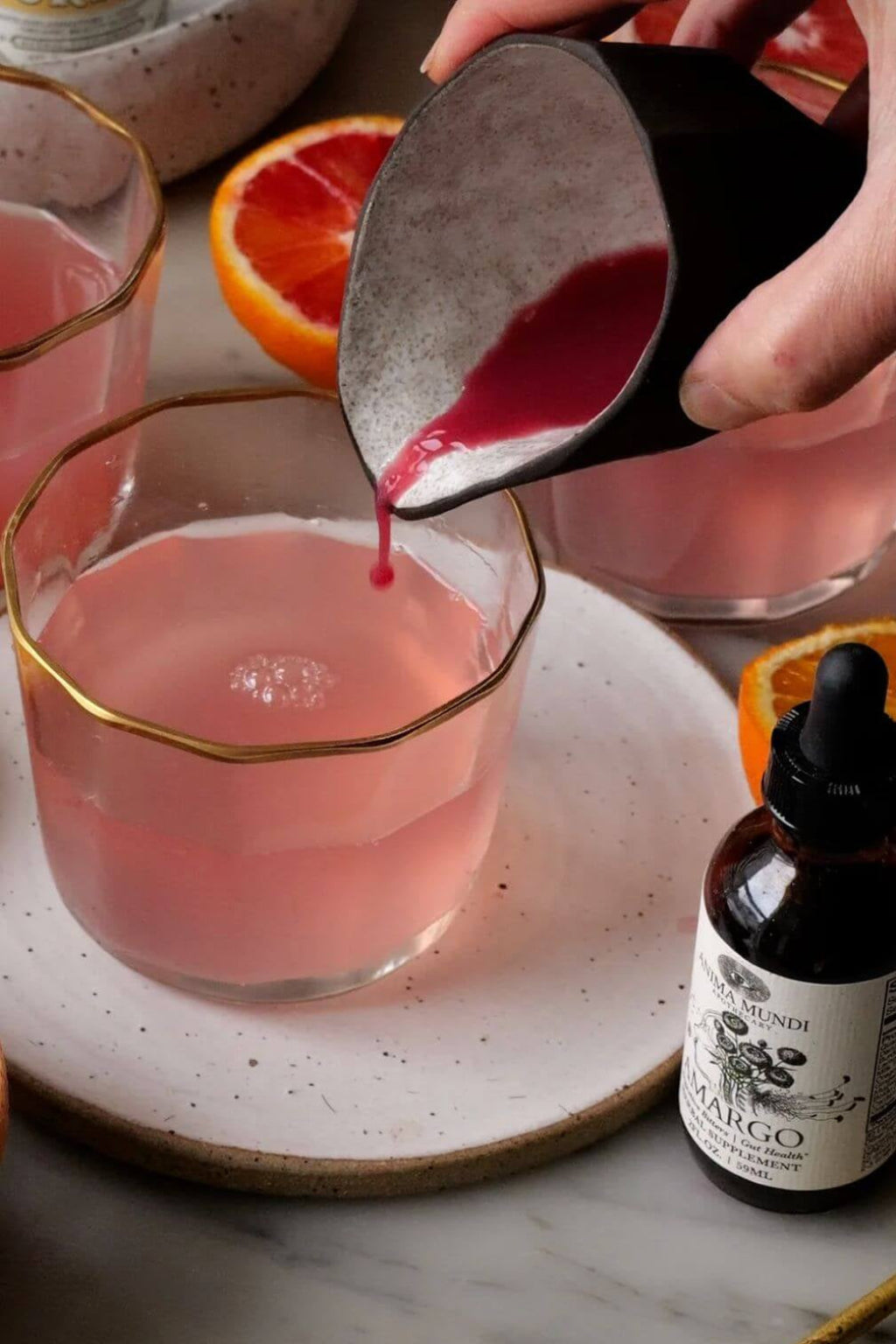 A hand pouring a dark pink liquid from a black ceramic pitcher into a pink drink in a gold-rimmed glass, set on a speckled white plate with orange slices and a small dropper bottle of Amargo Bitters nearby.