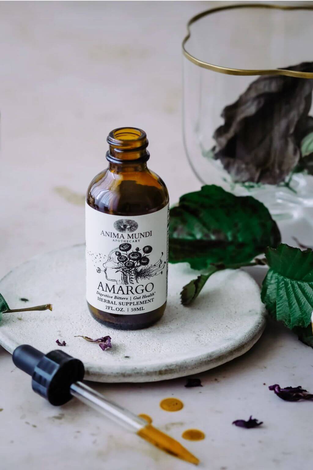 An amber glass dropper bottle labeled 'Anima Mundi Amargo' herbal supplement, styled on a light stone surface with green leaves, dried botanicals, and a glass container in the background.