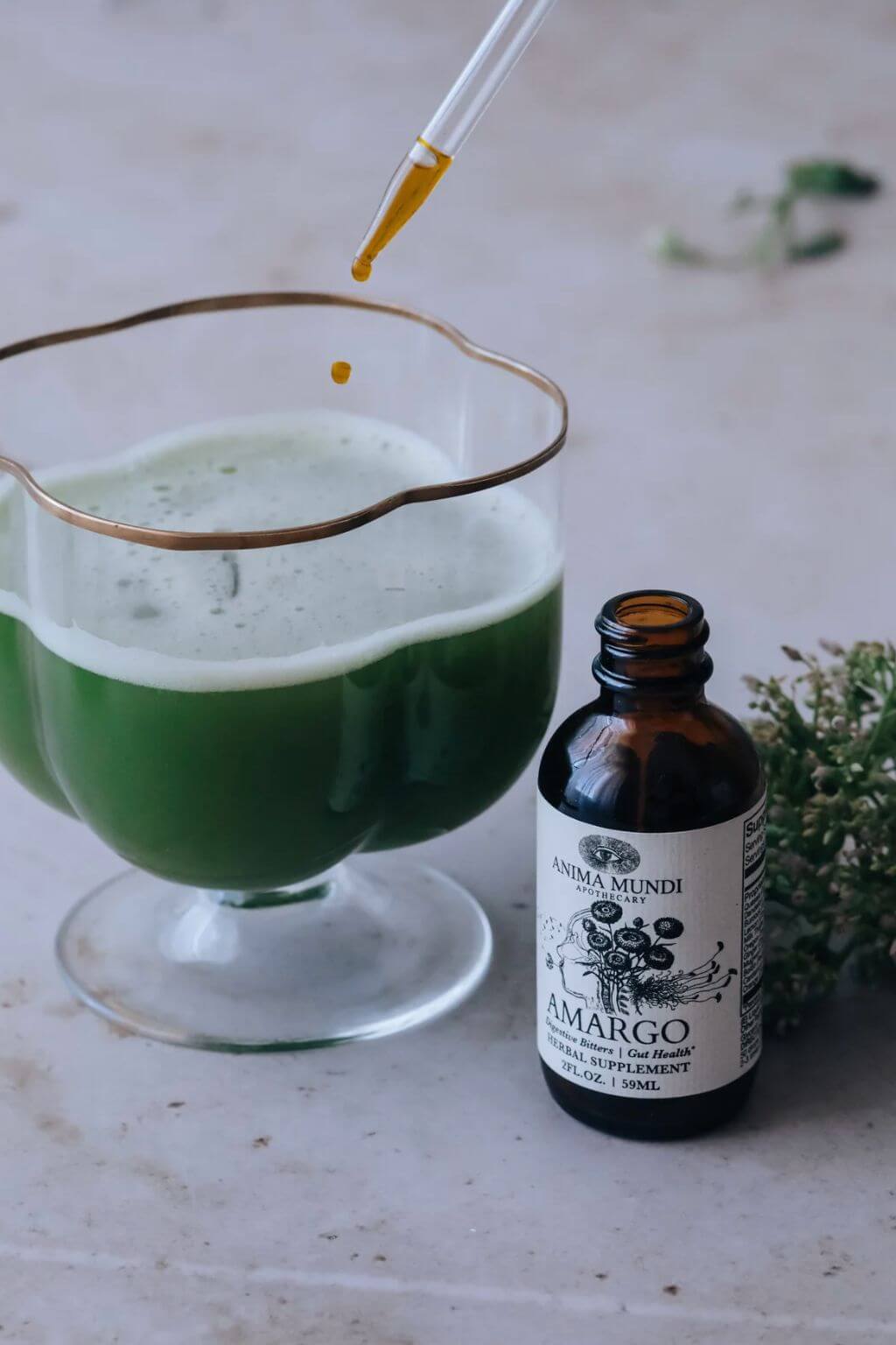 A decorative glass filled with green juice with a glass dropper above it, next to an amber bottle labeled Anna Mundi Amargo Bitters, on a light stone surface with a sprig of herbs.