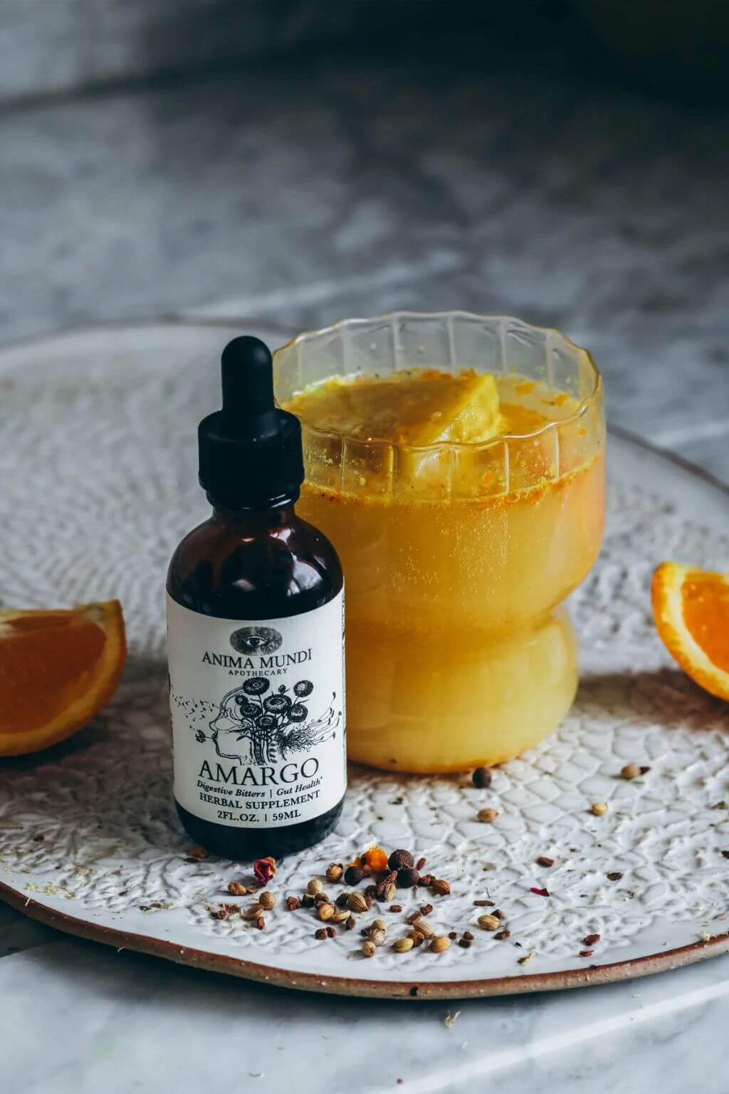 A small amber dropper bottle labeled Amargo by Anima Mundi stands beside a glass of orange-yellow drink on a textured white tray, with orange slices and peppercorns around it.