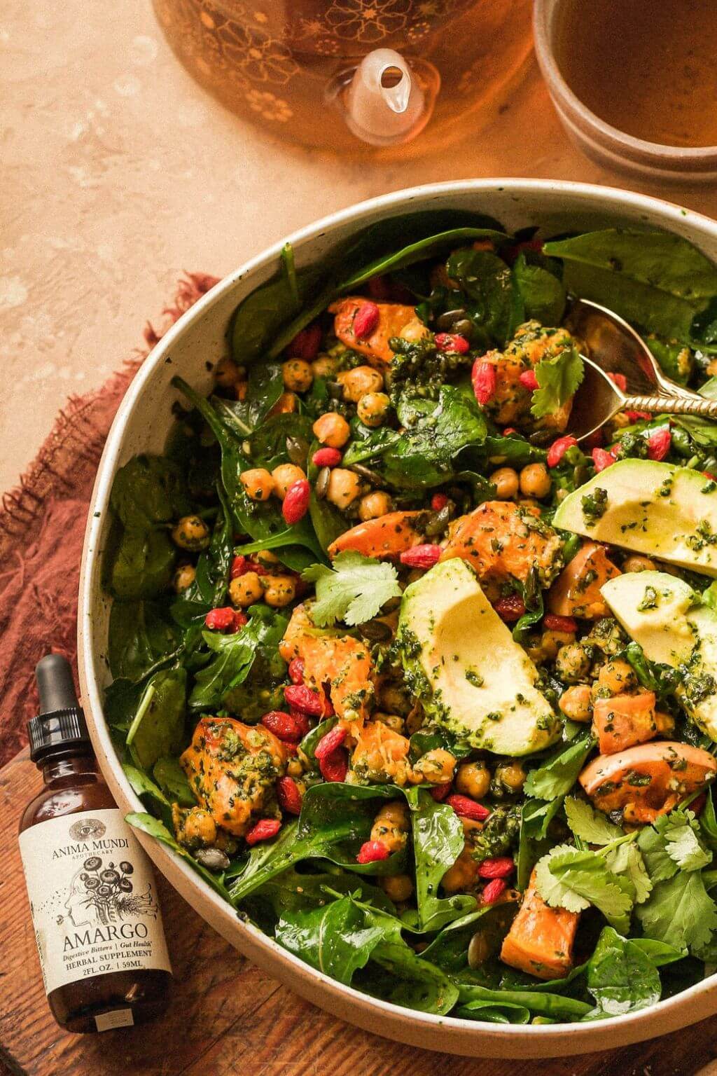 A large bowl filled with a green salad containing spinach, avocado, chickpeas, pomegranate, and grilled chicken, served on a rustic wooden table with a small bottle of Anima Mundi Amargo Bitters nearby.