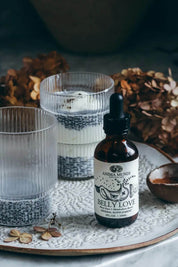 Anima Mundi Belly Love herbal supplement dropper bottle on a decorative tray, styled with two ribbed glass cups containing chia seeds and dried flowers in the background.
