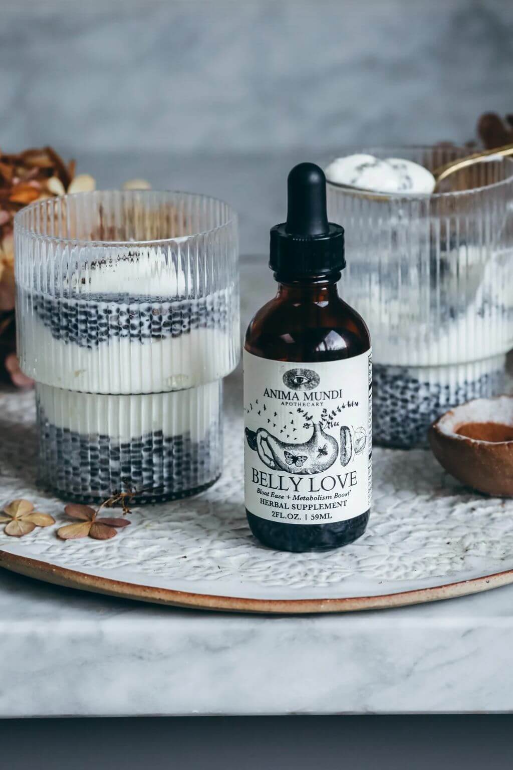 Anma Mundi Belly Love herbal supplement tincture in an amber glass dropper bottle, styled on a marble tray with layered chia seed and coconut drinks in ribbed glasses.