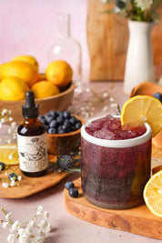 A styled tabletop scene with a small amber dropper bottle labeled Anima Mundi Belly Love beside a glass of dark berry drink with ice, lemon slices, blueberries, and flowers on a soft pink surface.