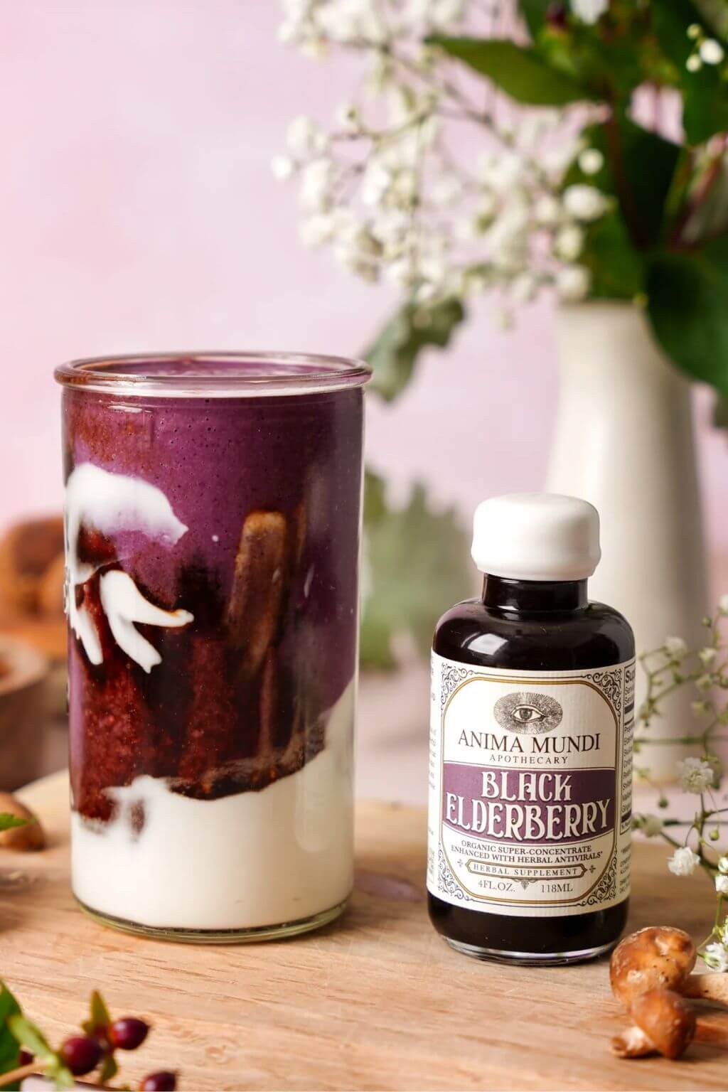 A bottle of Anima Mundi Black Elderberry syrup stands next to a glass of layered dark purple and white drink on a wooden surface, with flowers and ginger in the background.
