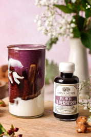 A bottle of Anima Mundi Black Elderberry syrup stands next to a glass of layered dark purple and white drink on a wooden surface, with flowers and ginger in the background.