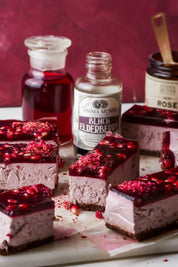 Several square slices of layered berry cheesecake with a dark berry gel topping and dried berry flakes are arranged on parchment paper. Two bottles and a jar of Anima Mundi ingredients, including Black Elderberry syrup and Rose Powder, stand behind the dessert on a dark burgundy background.