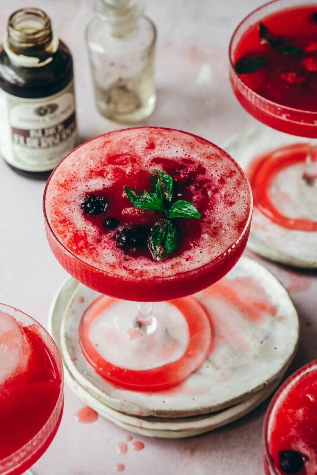 A frothy red cocktail in a coupe glass topped with mint leaves and berries, styled on a light surface with stacked plates, other red drinks, with a bottle of Black Elderberry Syrup in the background.
