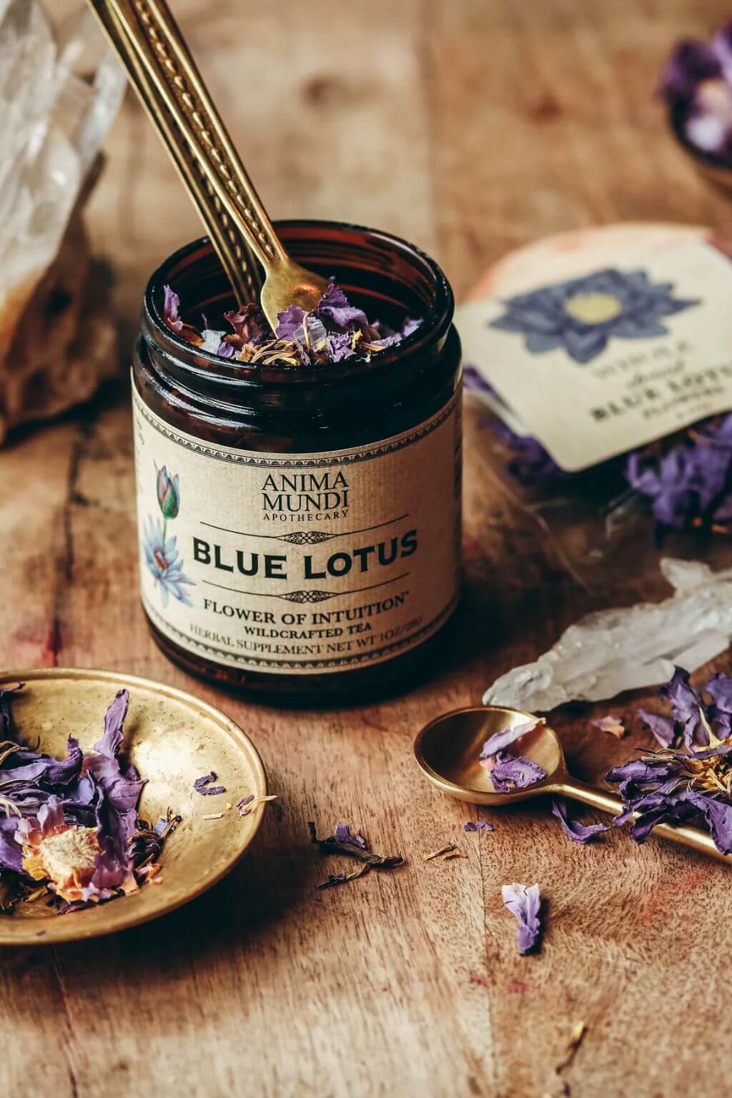 A jar of Anima Mundi Blue Lotus flower of intention tea sits on a rustic wooden table, surrounded by dried blue lotus petals, a brass spoon.