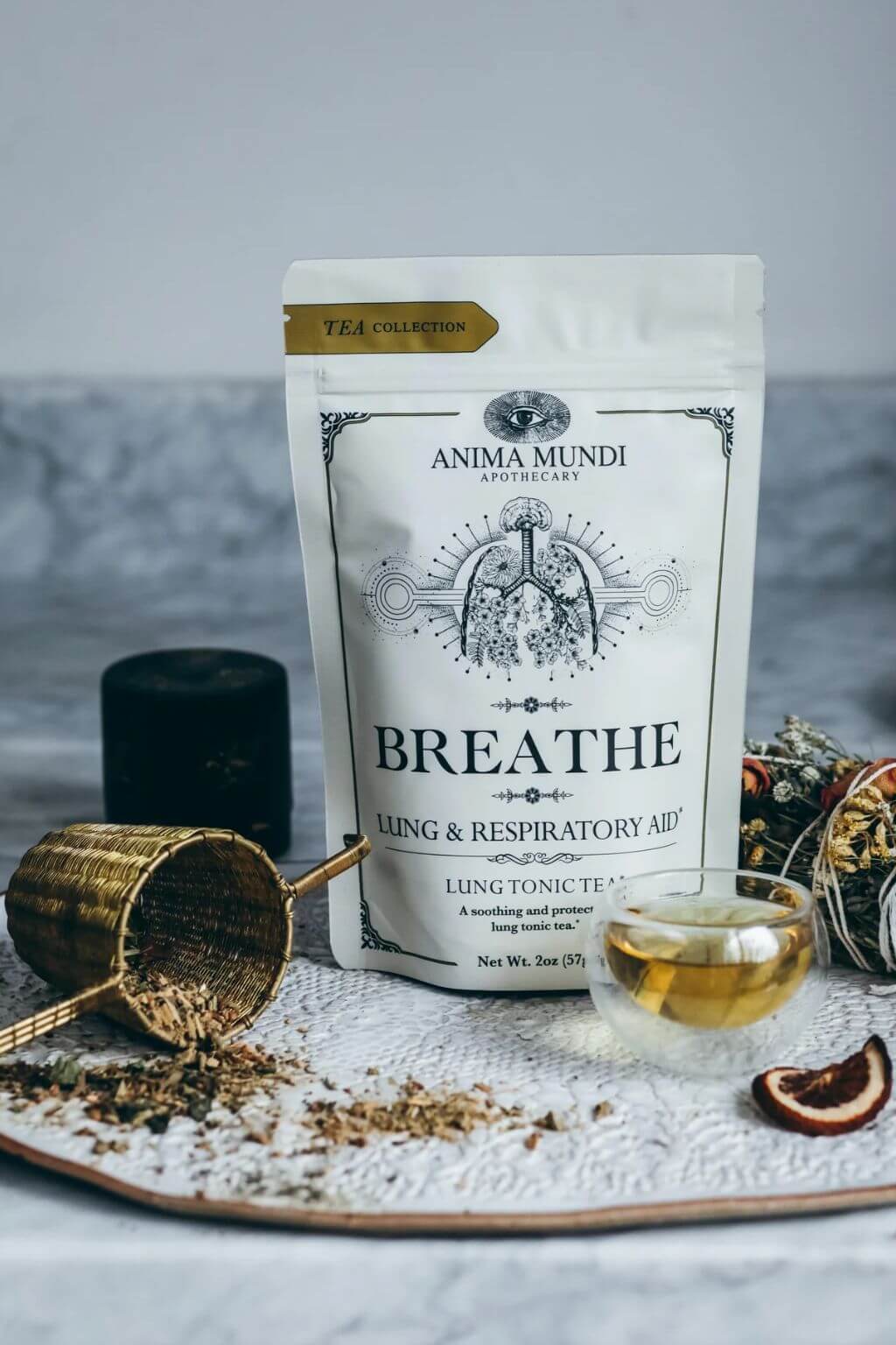 Anima Mundi 'Breathe' lung tonic tea in a white stand-up pouch, styled on a marble surface with loose tea, a small glass of golden tea, dried citrus slices, and a brass scoop.
