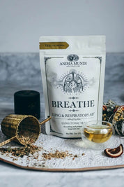Anima Mundi 'Breathe' lung tonic tea in a white stand-up pouch, styled on a marble surface with loose tea, a small glass of golden tea, dried citrus slices, and a brass scoop.