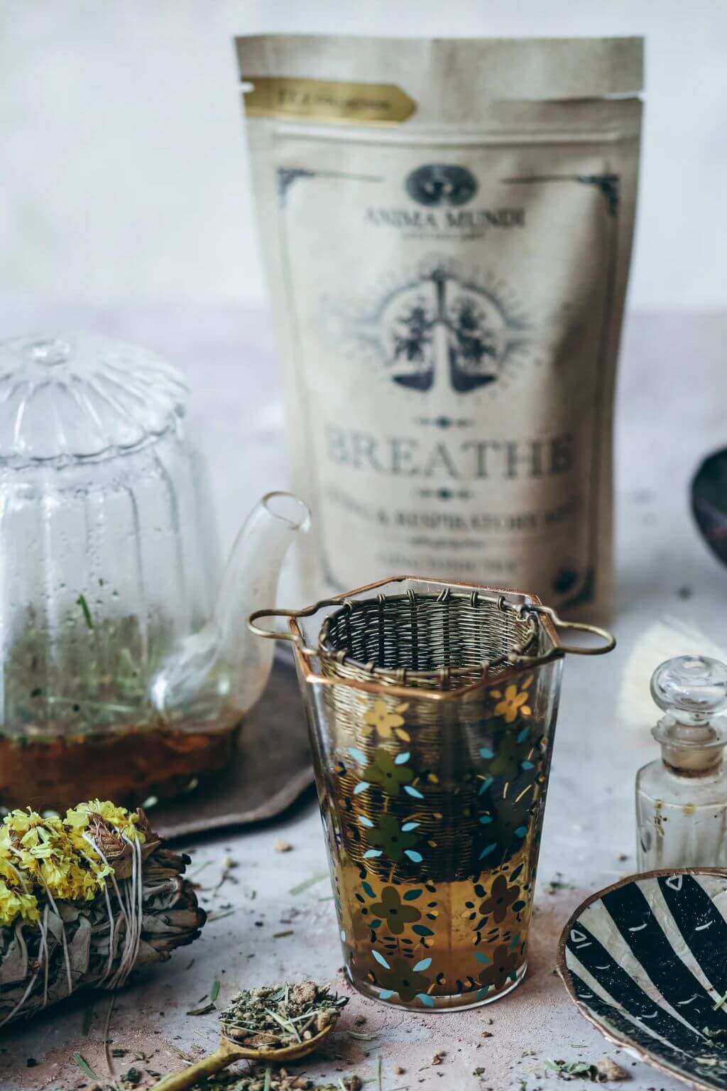Anima Mundi Breathe herbal tea pouch standing behind a woven brass tea infuser, with a glass teapot, loose herbs, and a small glass bottle on a light surface.