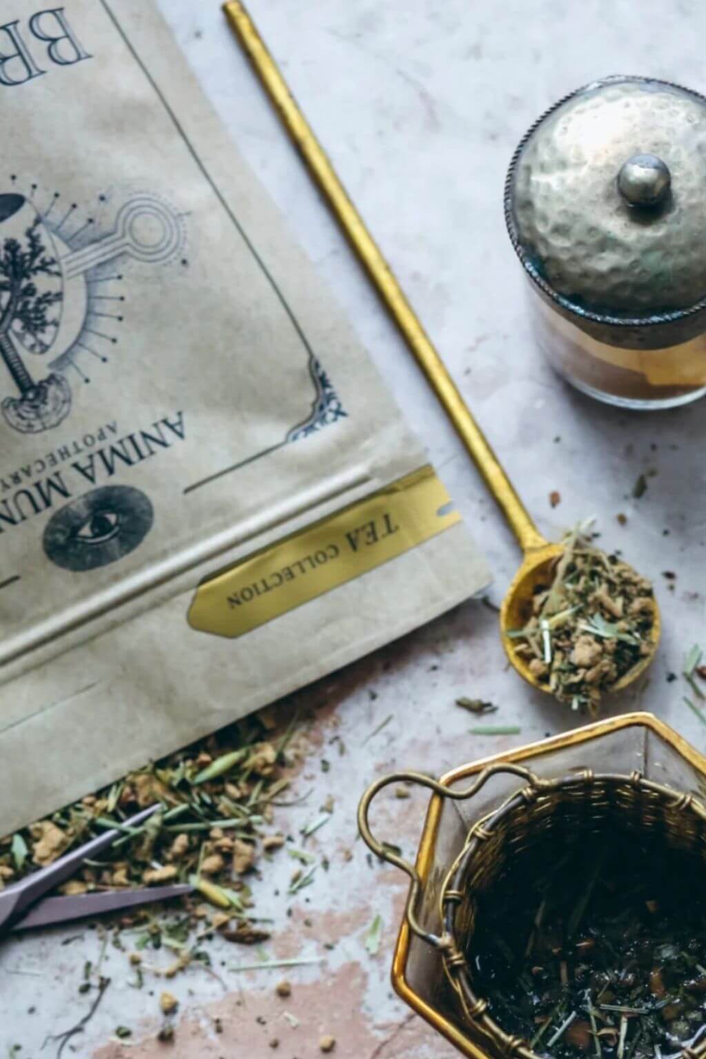 An open pouch labeled 'ANIMA MUNDI TEA COLLECTION' lies on a light stone surface with loose dried tea visible around it. A gold spoon holds some of the tea blend, next to a small hammered metal lidded container and a hexagonal metal infuser basket.