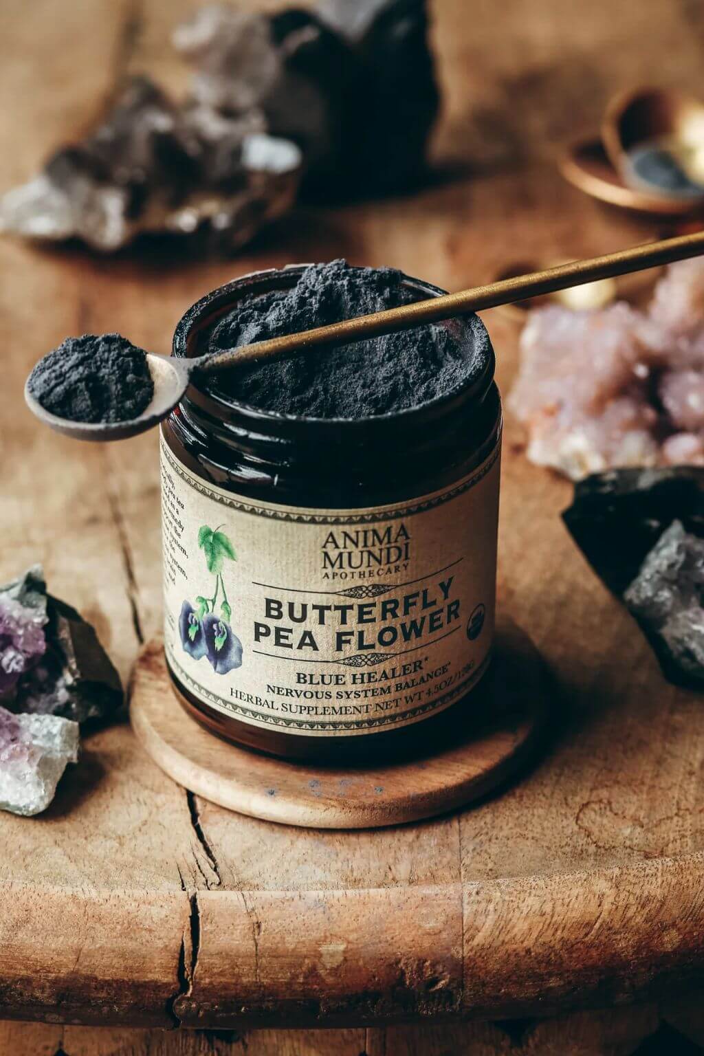 A jar of Anima Mundi Butterfly Pea Flower herbal supplement powder sits on a wooden coaster with a spoonful of the dark powder resting on the lid. The scene is styled with crystals and a rustic wooden background.