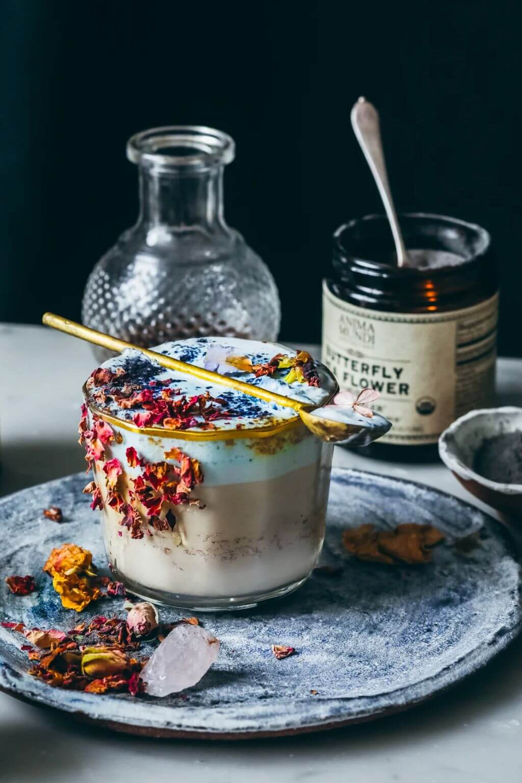 A styled product shot of a layered dessert in a clear glass cup, topped with dried flowers and a brass spoon, set on a gray ceramic plate. In the background, a jar labeled Butterfly Flower and a small glass bottle are visible against a dark backdrop.