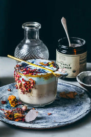 A styled product shot of a layered dessert in a clear glass cup, topped with dried flowers and a brass spoon, set on a gray ceramic plate. In the background, a jar labeled Butterfly Flower and a small glass bottle are visible against a dark backdrop.