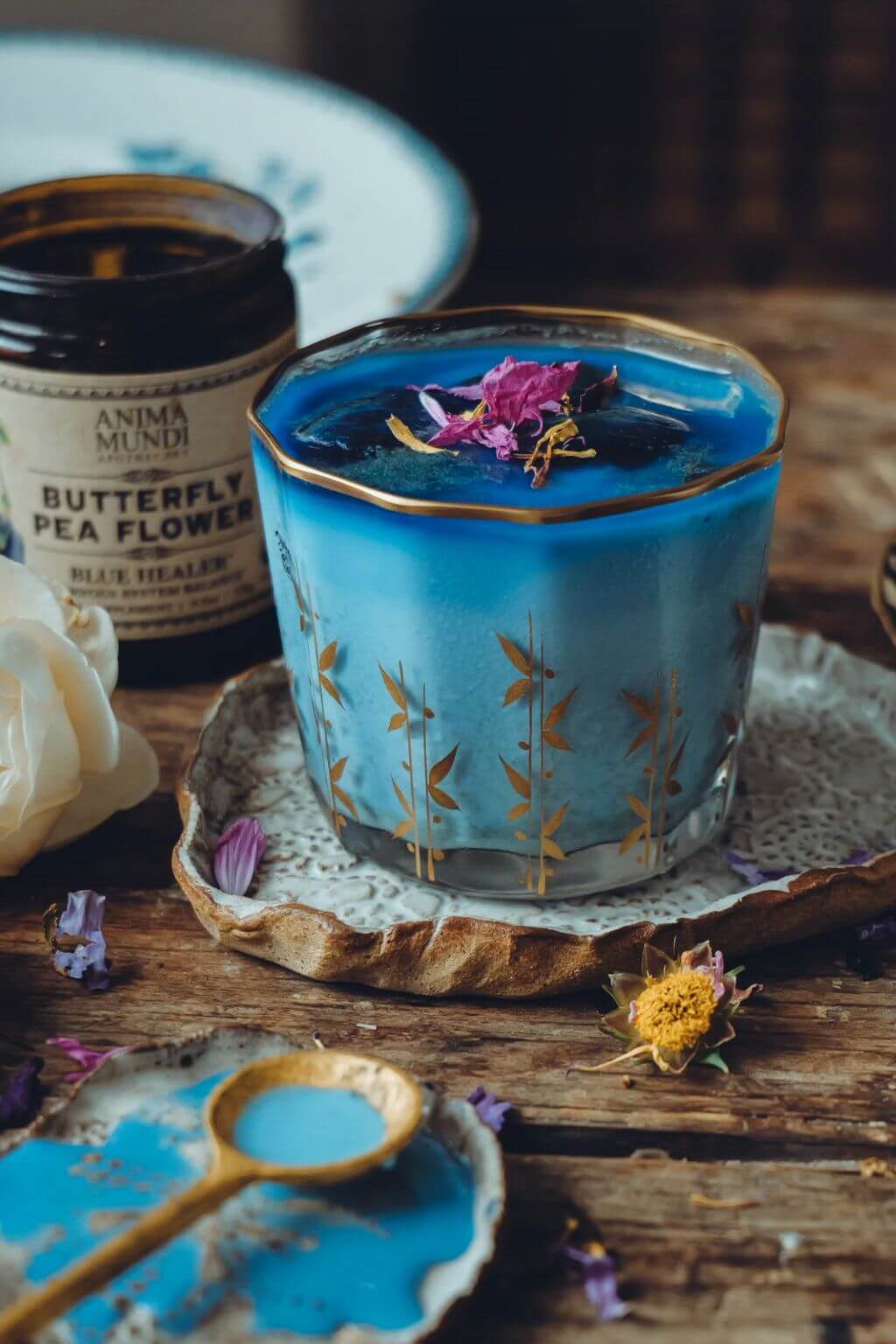 A vintage glass with gold leaf patterns filled with a blue beverage with a dried flower on top, styled on a rustic wooden table next to a jar of Anima Mundi Butterfly Pea Flower wax melt.