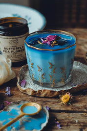 A vintage glass with gold leaf patterns filled with a blue beverage with a dried flower on top, styled on a rustic wooden table next to a jar of Anima Mundi Butterfly Pea Flower wax melt.