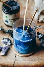 A glass mug filled with a vivid blue beverage sits on a wooden coaster on a rustic wooden table, with a brass straw inserted. A jar labeled 'Butterfly Pea Powder' by Anima Mundi is visible in the background along with crystals and greenery.