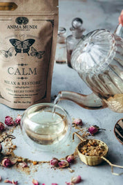 Anima Mundi Calm Relax & Restore herbal tea, shown with its kraft paper pouch, a glass teapot pouring tea, and a glass cup on a textured gray surface with dried rosebuds and a brass scoop.