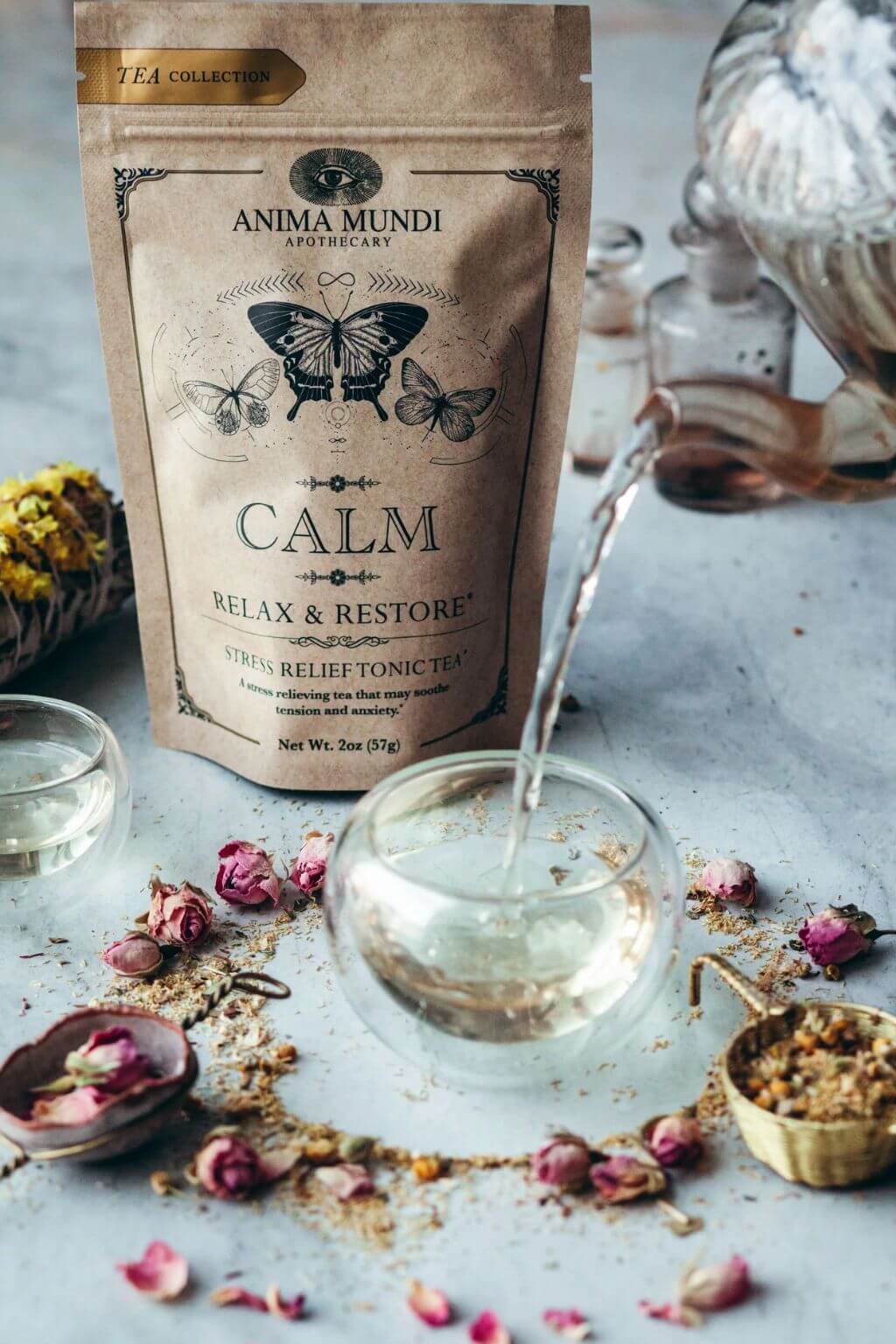 Anima Mundi Apothecary Calm Relax & Restore tea collection pouch, with tea being poured from a glass pitcher into a glass cup, surrounded by dried flowers and tea ingredients on a light gray surface.