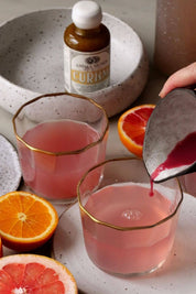 A hand pours a dark pink liquid from a metal pitcher into two clear lowball glasses with gold trim, set on a speckled white plate. A small bottle labeled Anima Mundi Curam is visible in the background, with orange and grapefruit slices around the glasses.