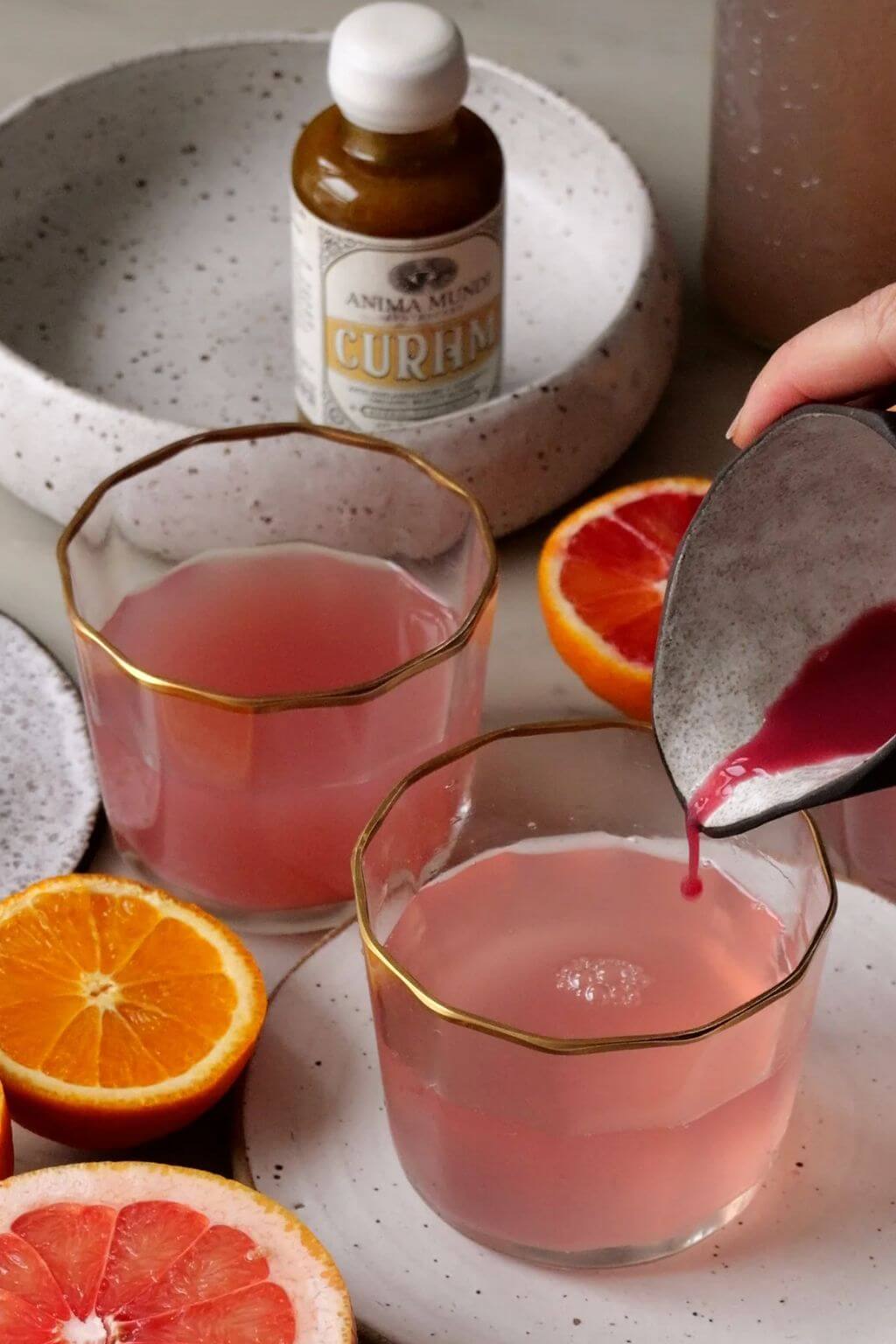 A hand pours a dark pink liquid from a metal pitcher into two clear lowball glasses with gold trim, set on a speckled white plate. A small bottle labeled Anima Mundi Curam is visible in the background, with orange and grapefruit slices around the glasses.