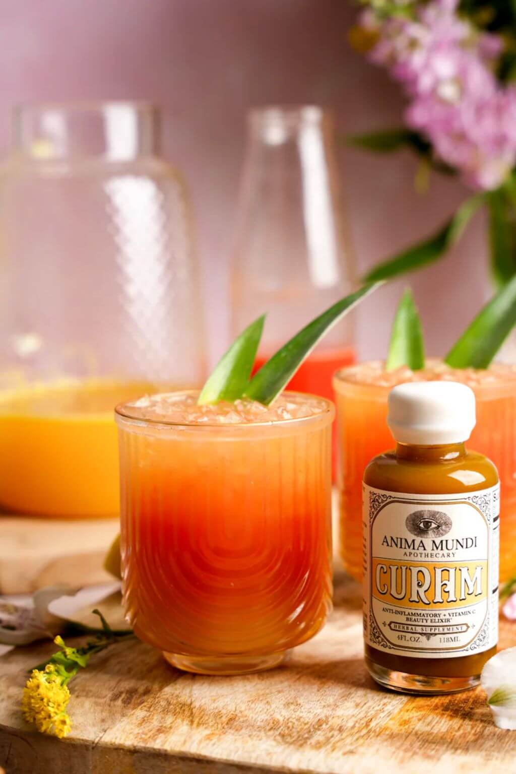 A small bottle of Anima Mundi Curam herbal syrup next to two glasses of orange-colored drink garnished with green leaves, set on a wooden board with a soft-focus background.