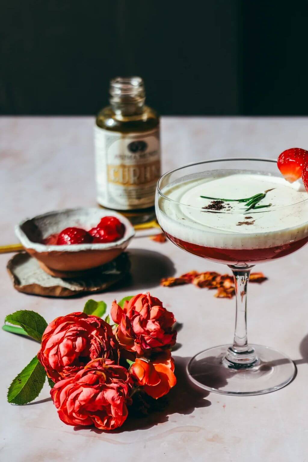 A styled cocktail scene on a pale stone surface with a stemmed glass containing a layered drink, a small bowl of red berries, a bottle of Curam in the background, and red flowers in the foreground.