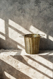 A small woven  brass infuser basket with two loop handles sits on a rough stone surface against a textured concrete wall, lit by strong diagonal shadows.