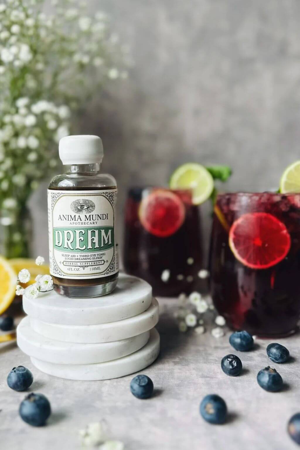 A small bottle of Anima Mundi 'Dream' Elixir displayed on a stack of white ceramic dishes, with two dark berry cocktails garnished with lime slices in the background.